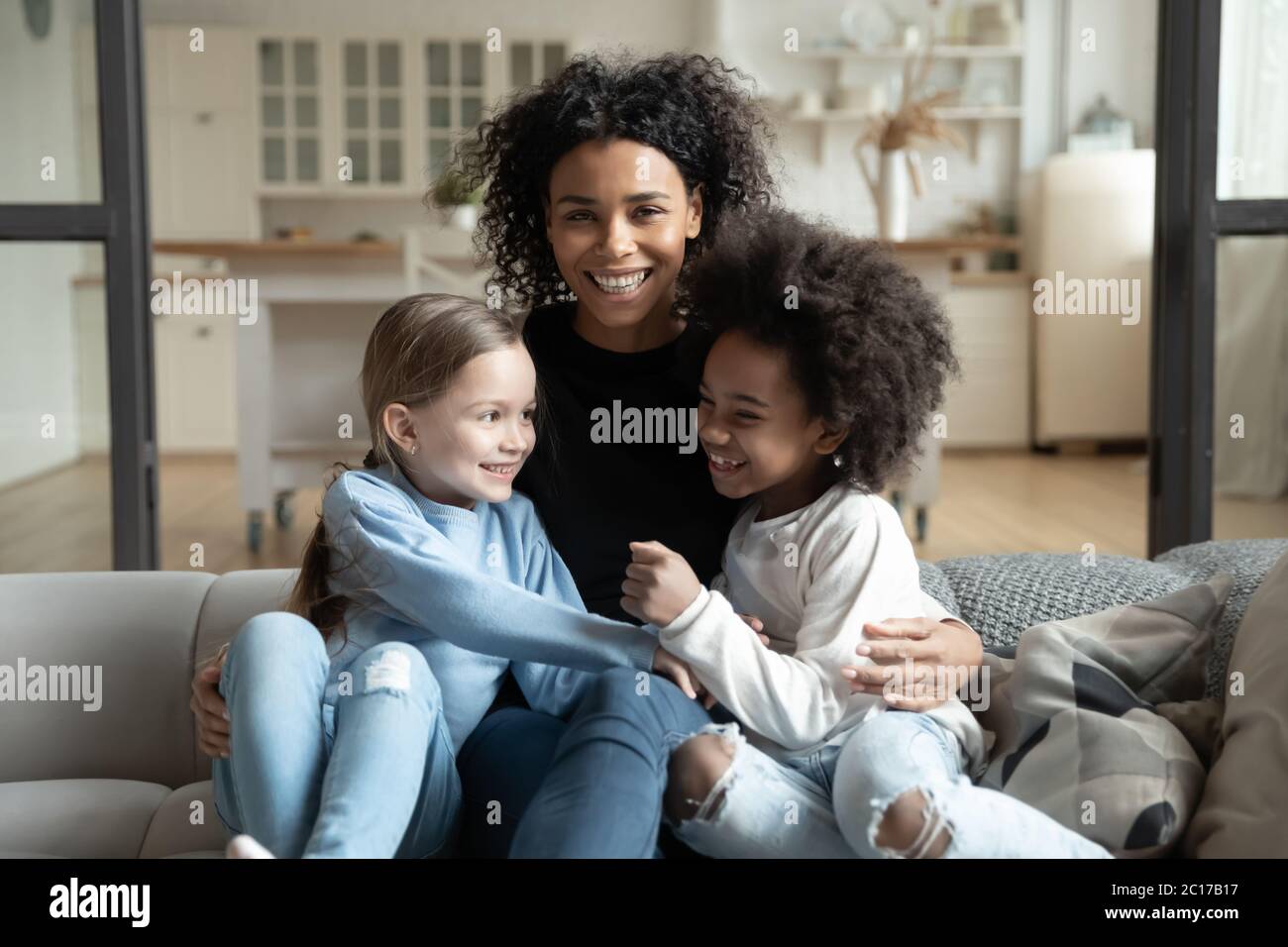 Cheerful African mother sitting on couch embraces multiracial daughters Stock Photo
