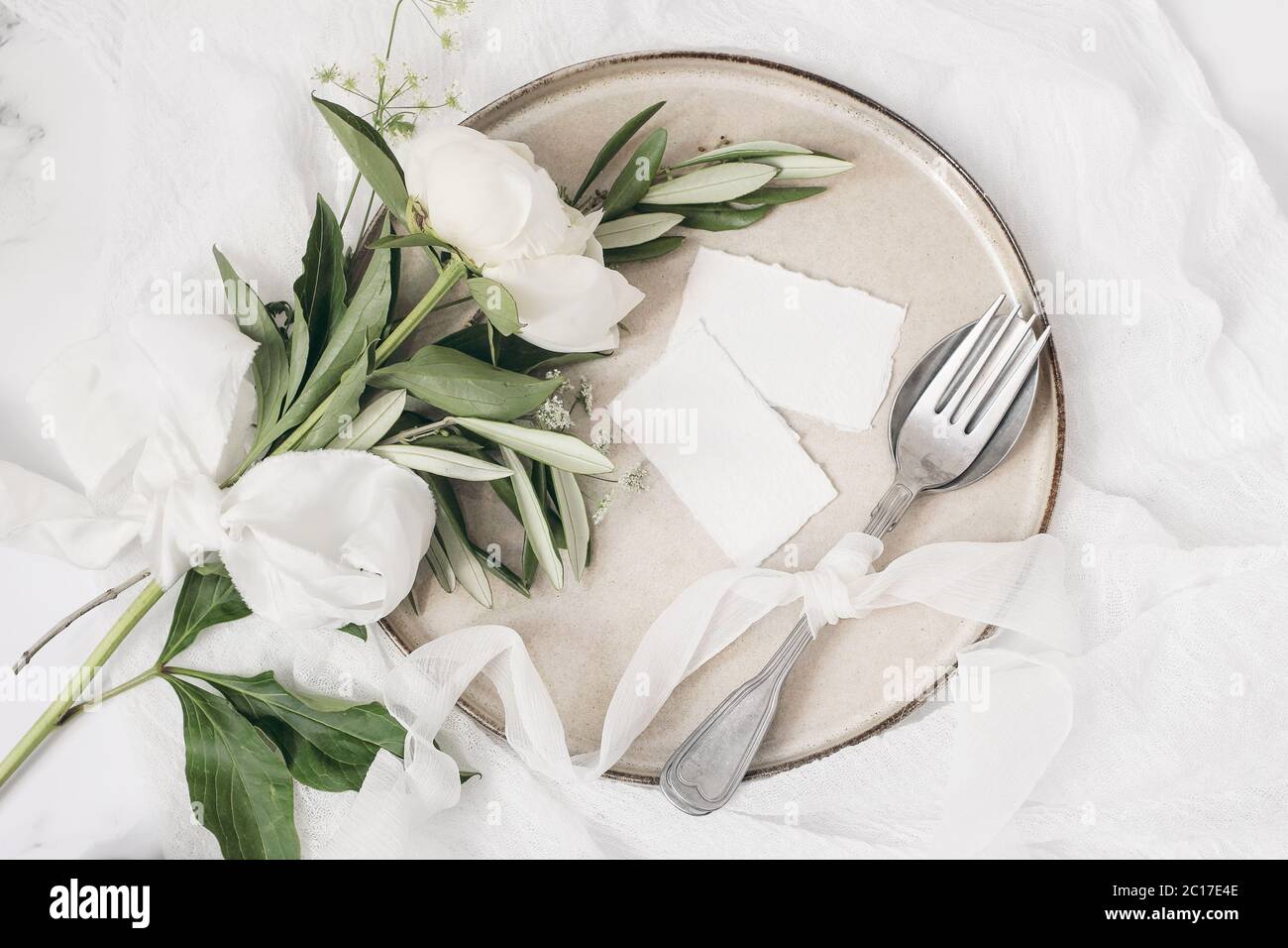 Festive summer wedding scene. Marble table setting with cutlery, olive branches, white peony flowers, stoneware plate and silk ribbon. Blank place Stock Photo