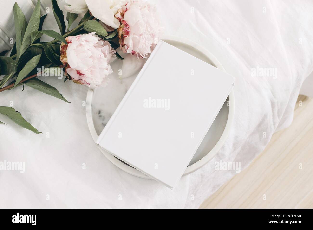 Wedding still life scene. White empty book cover mockup on marble tray. Pink peony flowers on white linen table cloth. Vintage feminine styled photo Stock Photo