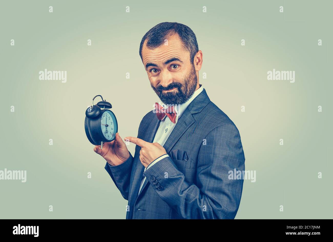 Mature man in suit showing an alarm clock. Businessman with an alarm clock in a hand looking at camera. Mixed race bearded model isolated on green yel Stock Photo