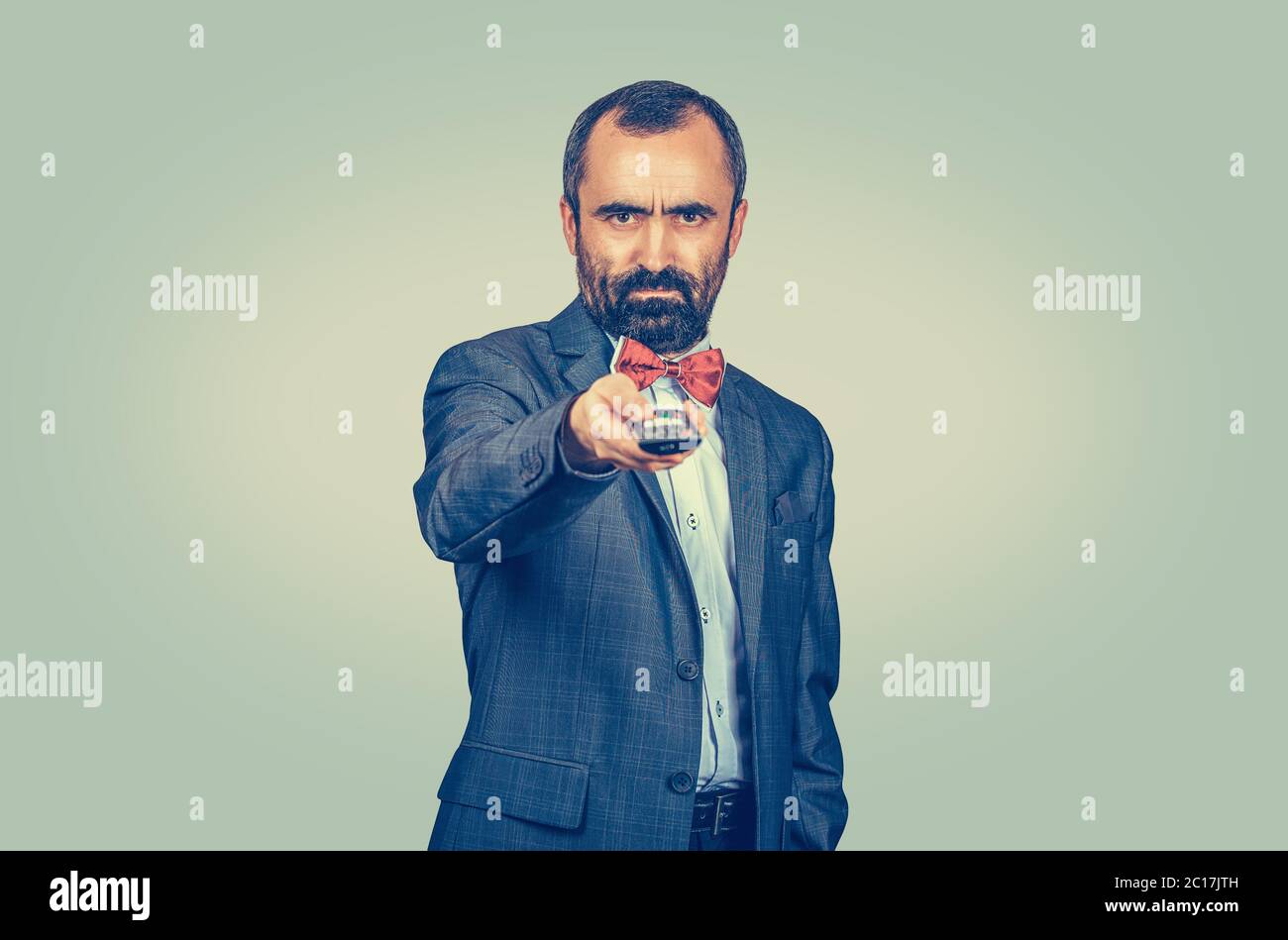 Adult displeased bearded man in elegant suit with red bow tie holding remote tv control. Mixed race bearded model isolated on green yellow uniform gra Stock Photo