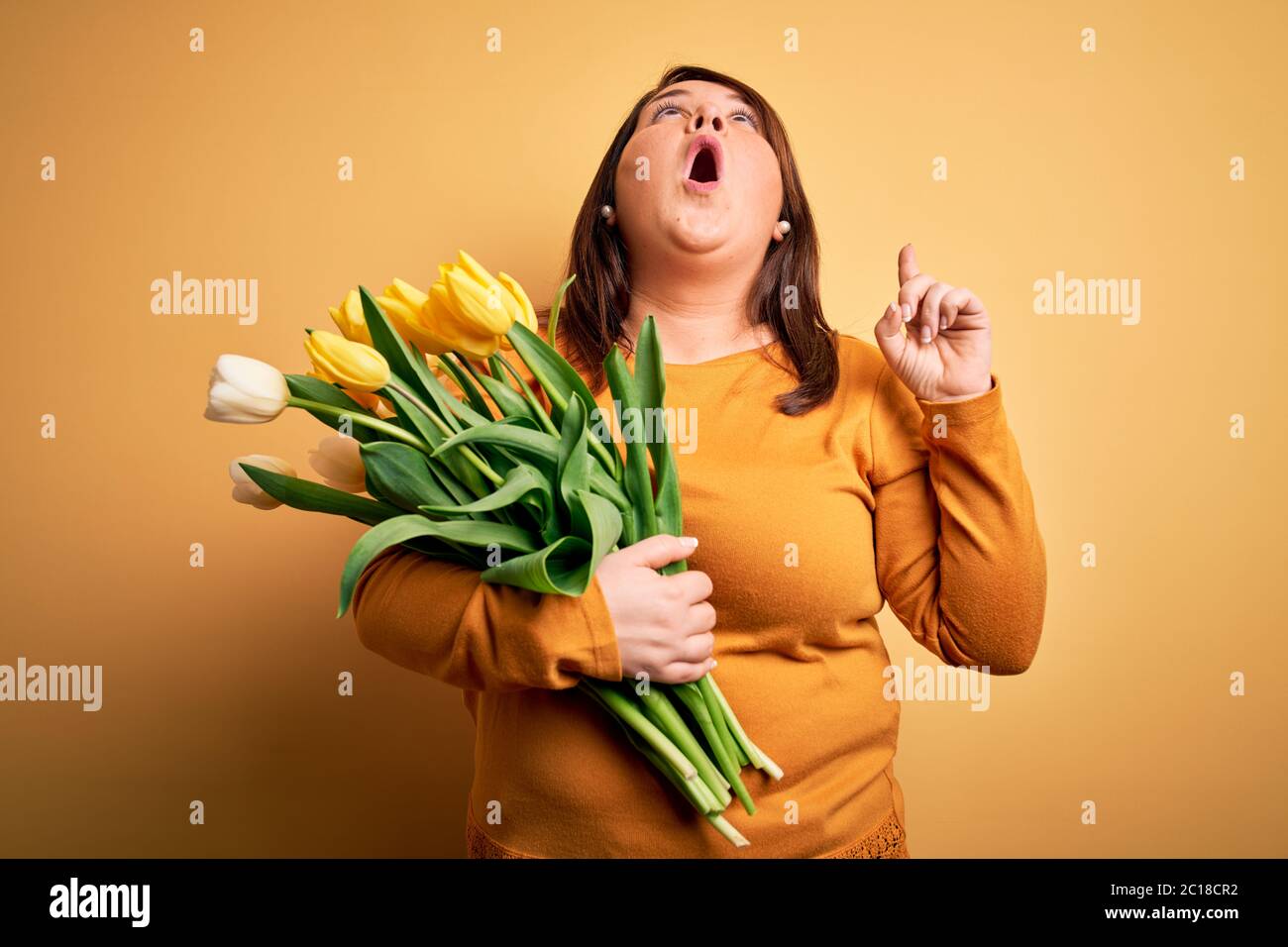 Beautiful plus size woman holding romantic bouquet of natural tulips flowers over yellow background amazed and surprised looking up and pointing with Stock Photo
