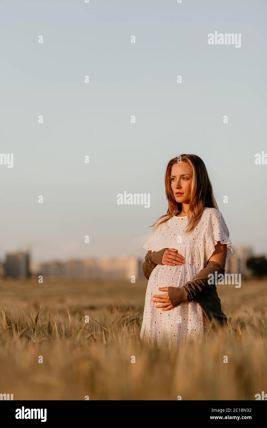 Young beautiful pregnant woman in white dress walking in the wheat orange field on a sunny summer day. Nature in the country. Miracle expectation Stock Photo