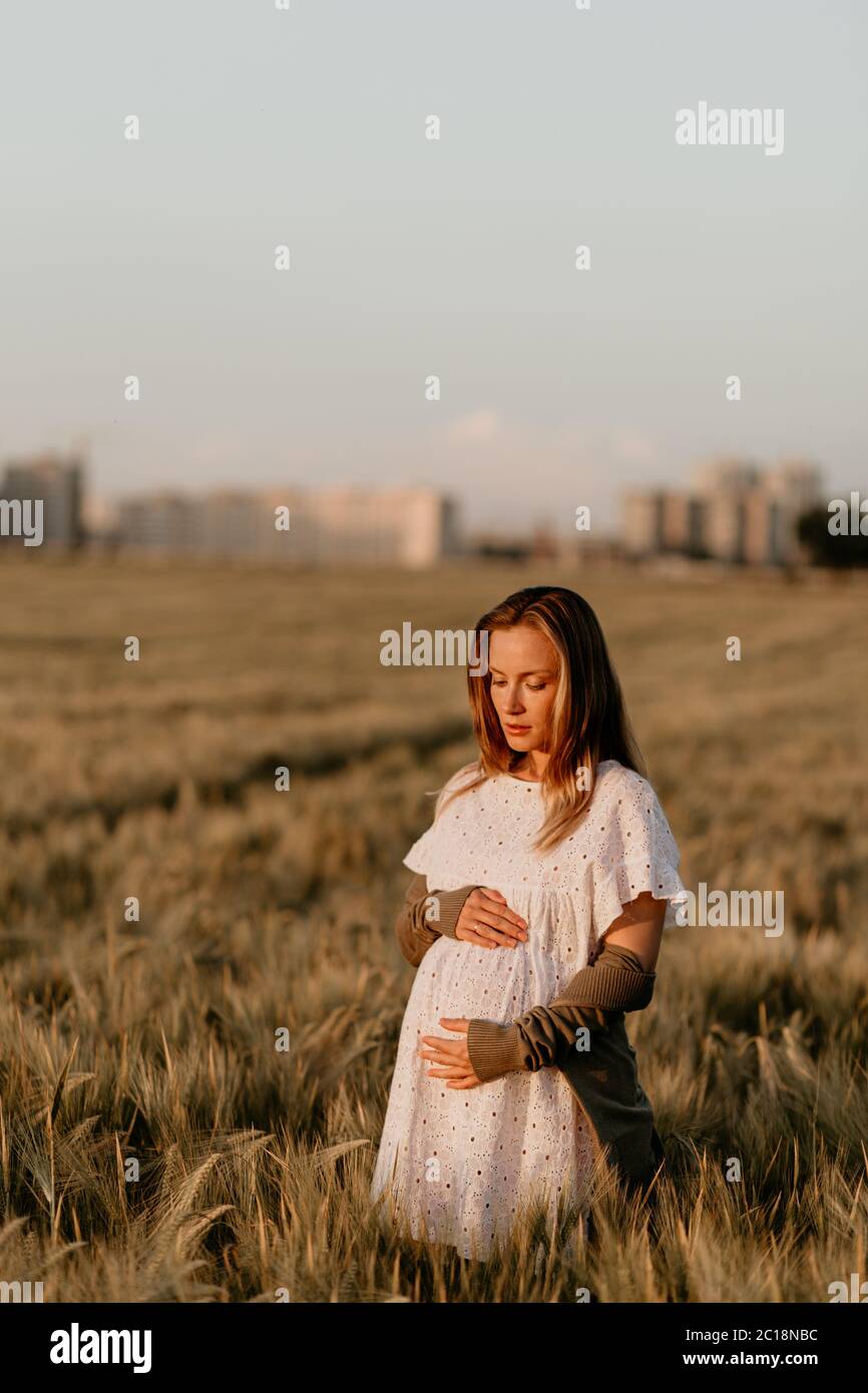 Young beautiful pregnant woman in white dress walking and touching her belly in the wheat orange field on a sunny summer day. Nature in the country Stock Photo