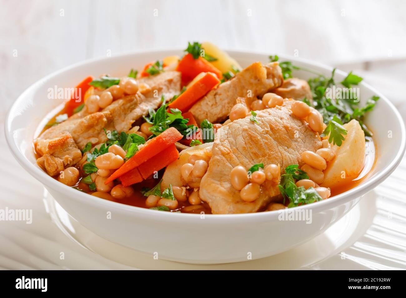 close-up of a portion of white bean, vegetables and pork stew in a white bowl on a rustic table, landscape view from above, Stock Photo