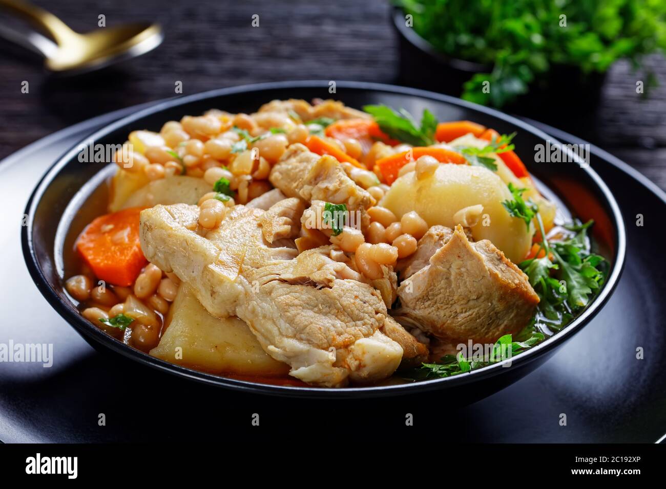 close-up of a portion of white bean, vegetables and pork steak stew in a black bowl on a dark wooden table Stock Photo