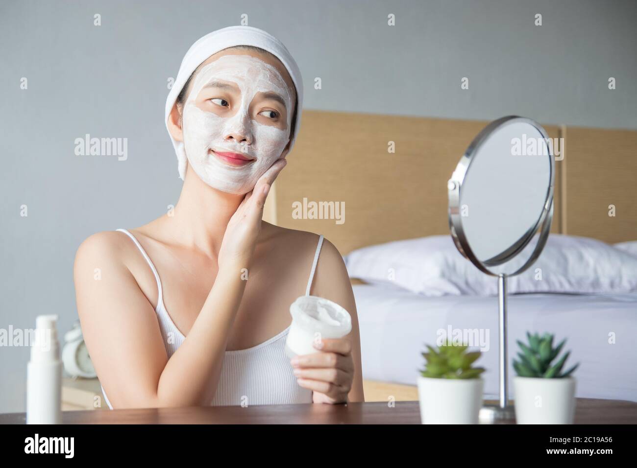 Asian woman applying moisturizer face scrub peeling white clay mask on skin, looking in mirror with smiley face using cream, touching face. Stock Photo