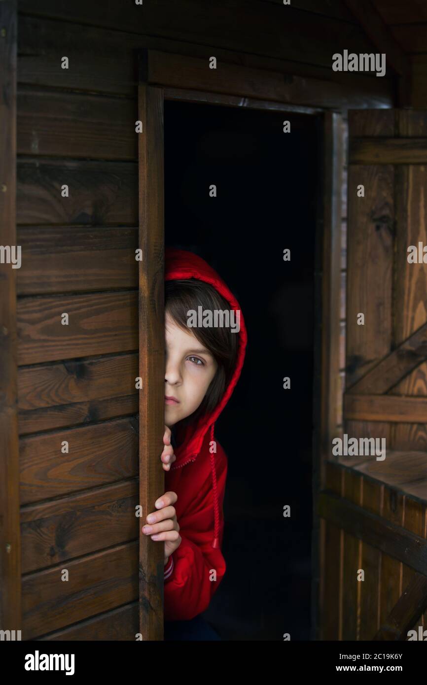 Preteen boy in red sweatshirt, hiding behind a wooden door, looking scared and sad Stock Photo