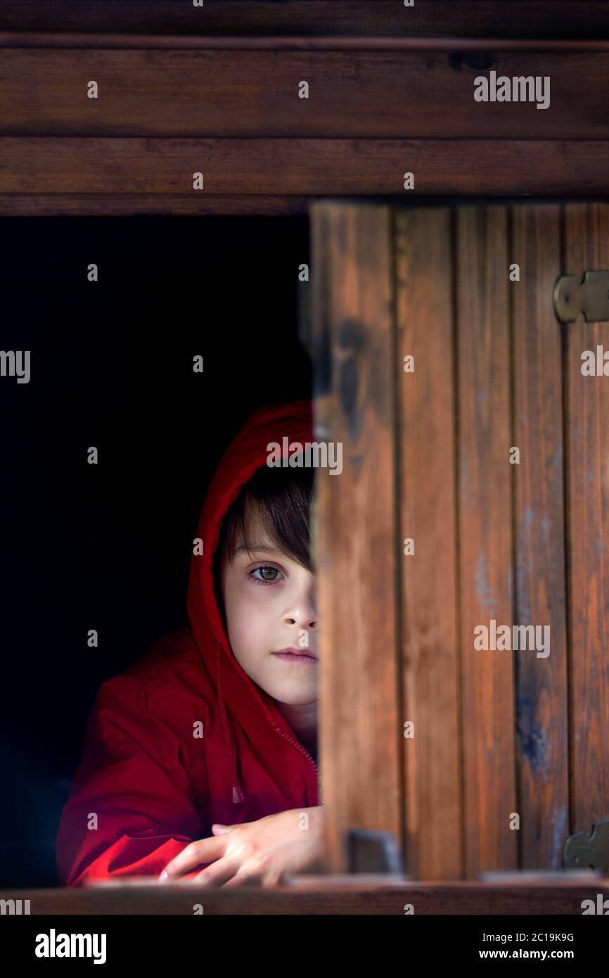 Preteen boy in red sweatshirt, hiding behind a wooden door, looking scared and sad Stock Photo