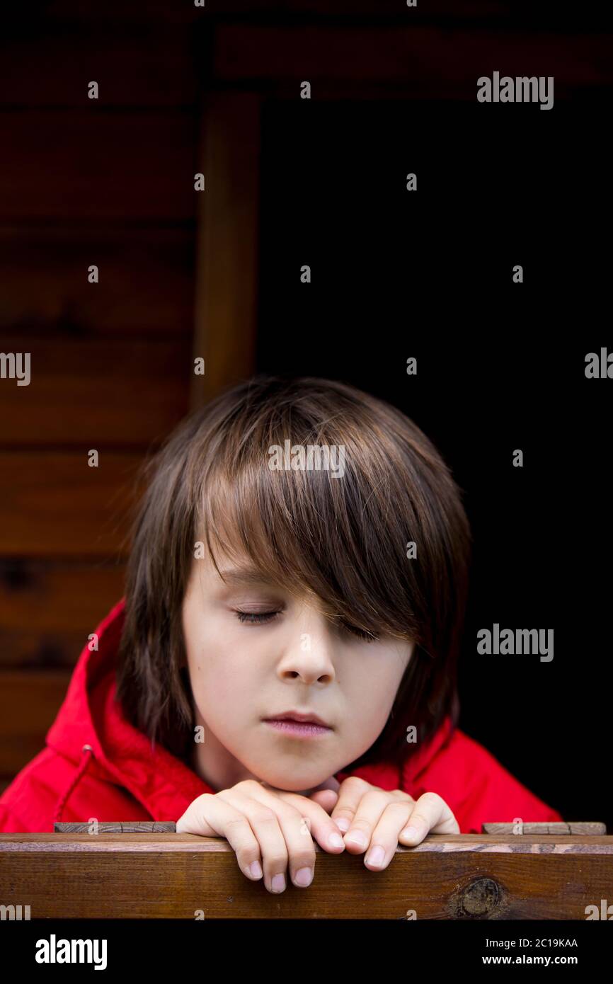 Preteen boy in red sweatshirt, hiding behind a wooden door, looking scared and sad Stock Photo