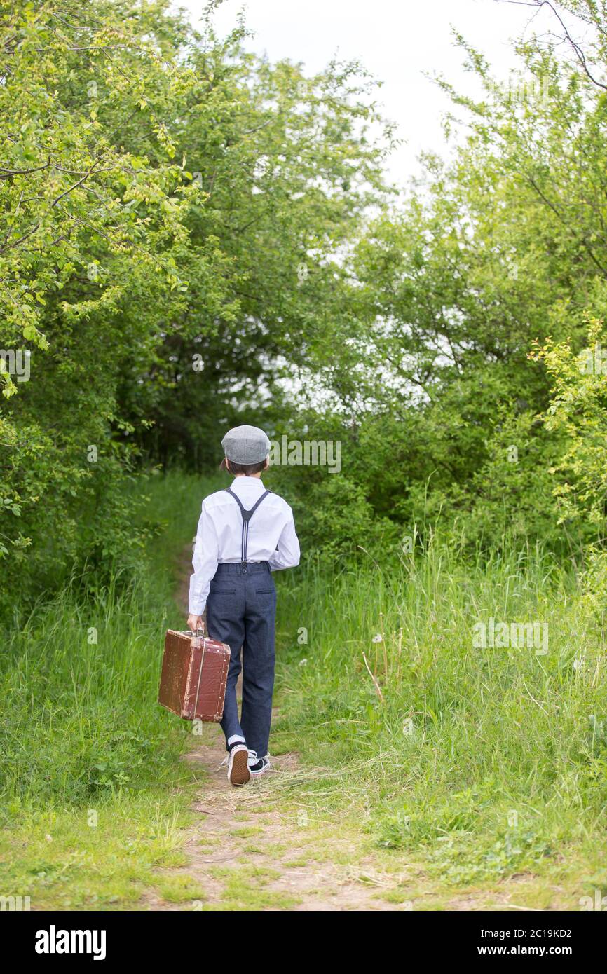 Sweet child in vintage clothing, hat, suspenders and white shirts, holding suitcase, running in the park, going to a holiday Stock Photo