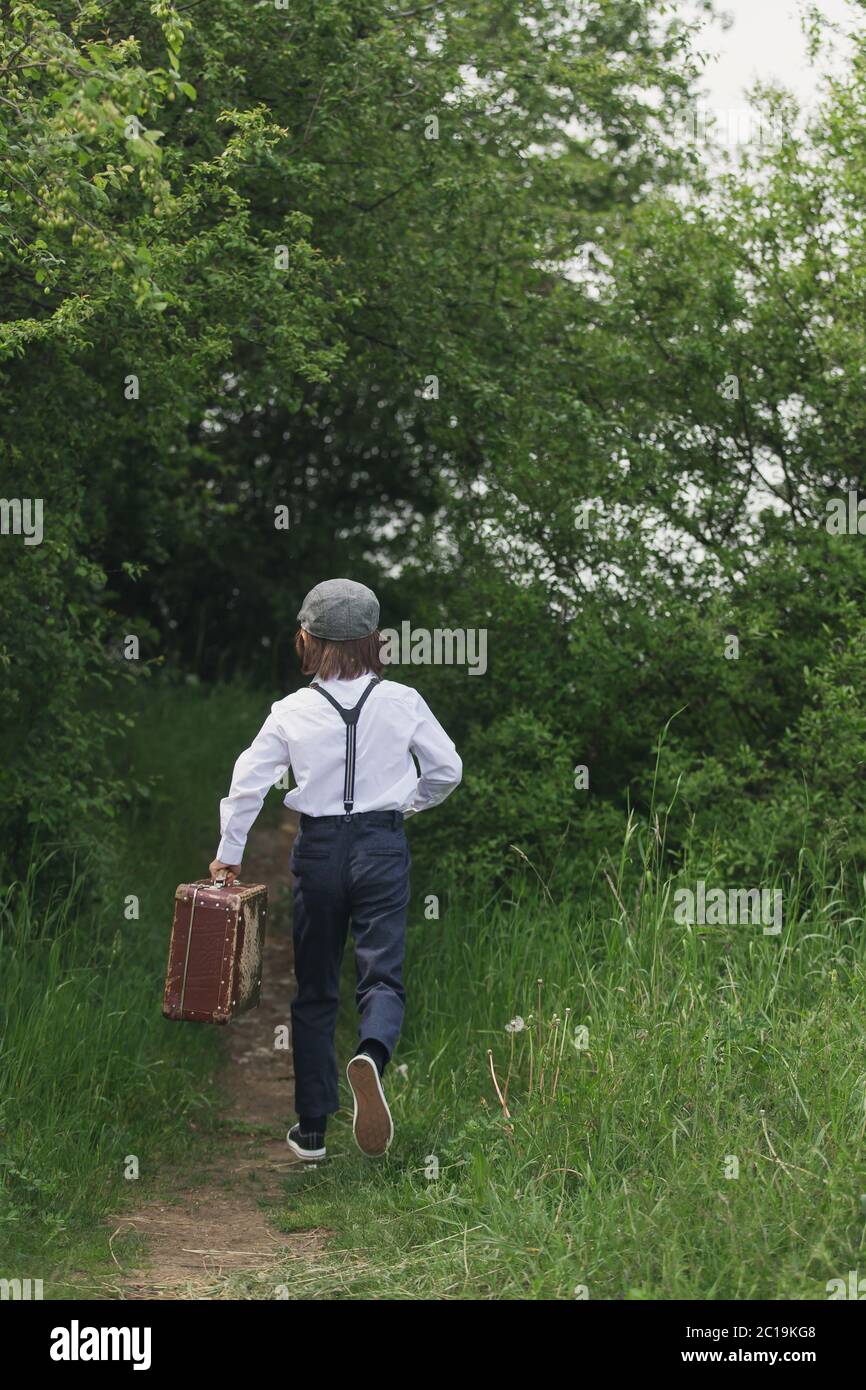 Sweet children in vintage clothing, hat, suspenders and white shirts, holding suitcase, running in the park, going to a holiday Stock Photo