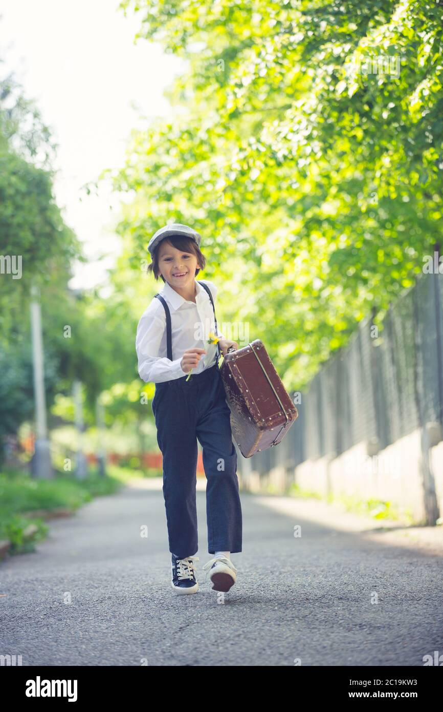 Sweet child in vintage clothing, hat, suspenders and white shirts, holding suitcase in the park, going to a holiday Stock Photo