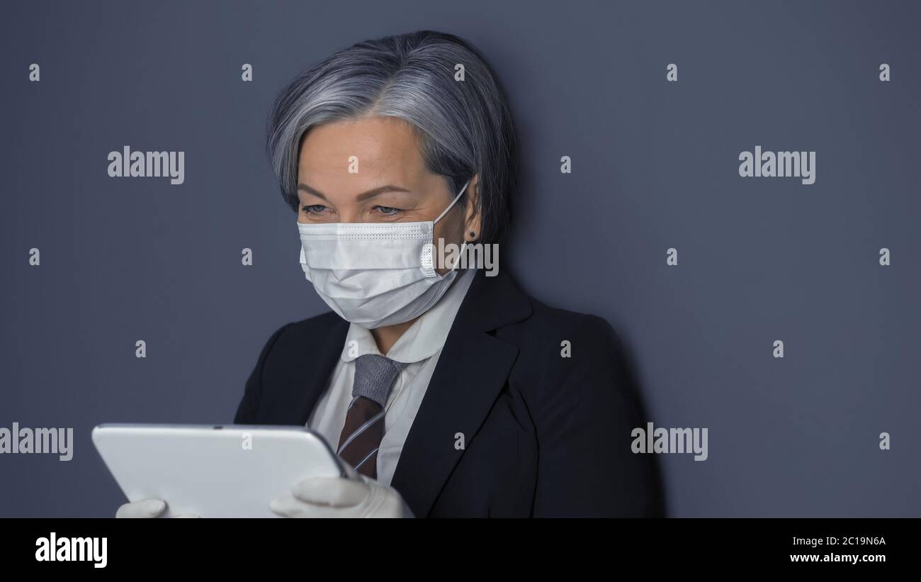 Mature businesswoman in protective mask using digital tablet. Gray haired woman reading news or working at tablet computer while standing on gray wall Stock Photo