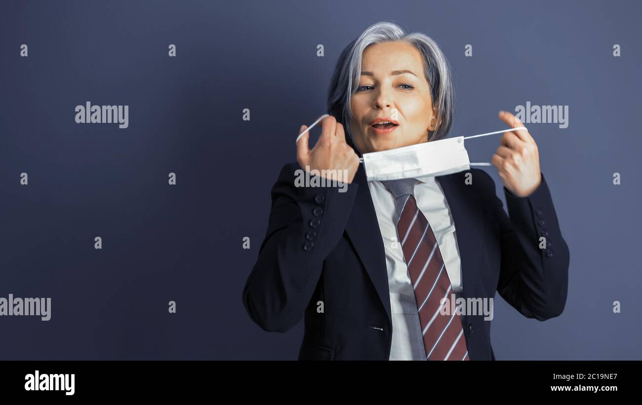 Business woman removes protective mask from her face. Focus on female face. Mature woman in formalwear on blue gray background. Copy space at left Stock Photo