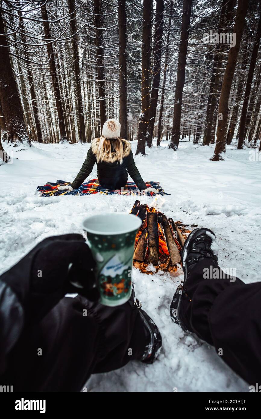 Young romantic couple having fun outdoors in the forest in winter season. Boyfriend sitting in the snow near the campfire and holding hot tea cup. Stock Photo
