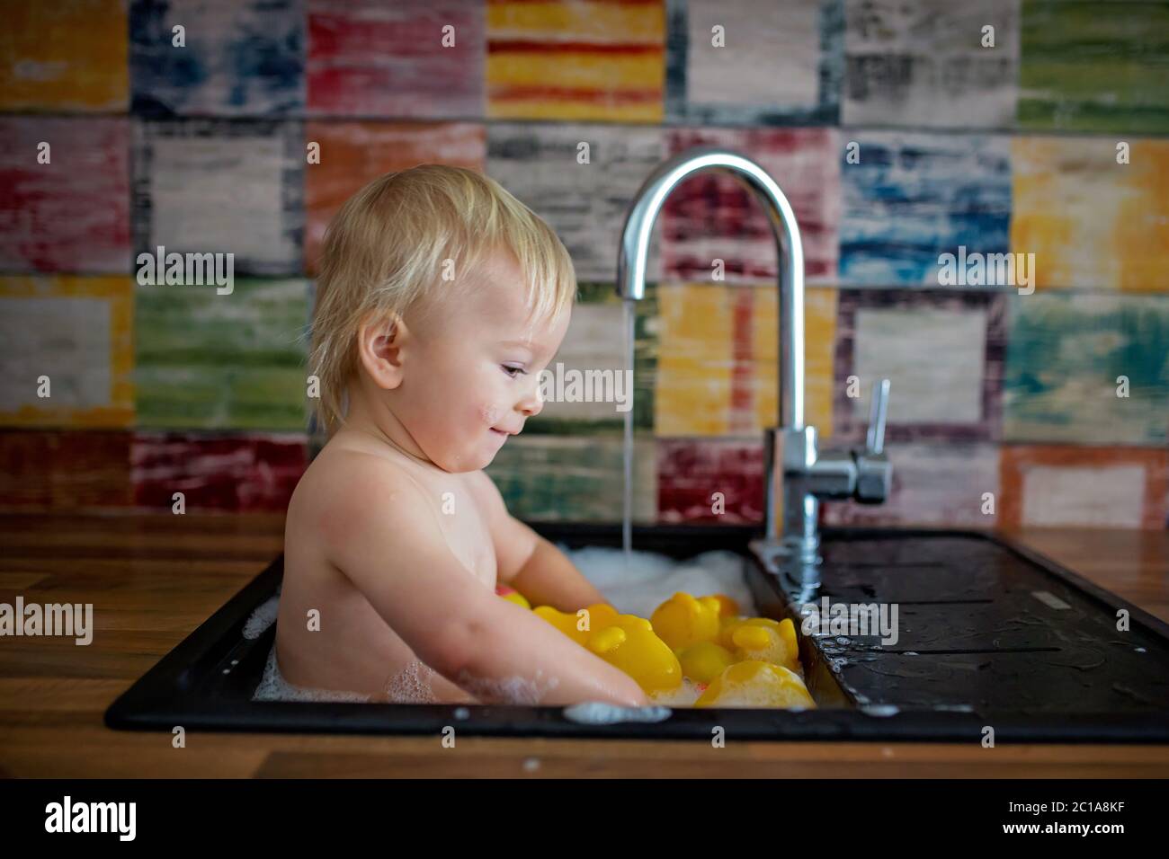 Cute smiling baby taking bath in kitchen sink. Child playing with foam