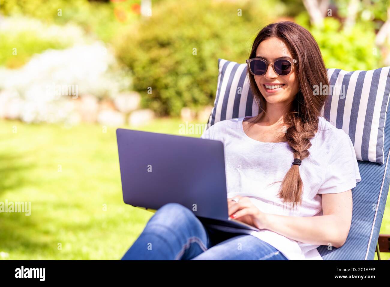 Shot of happy young woman sitting on sunbed in the garden and using her ...