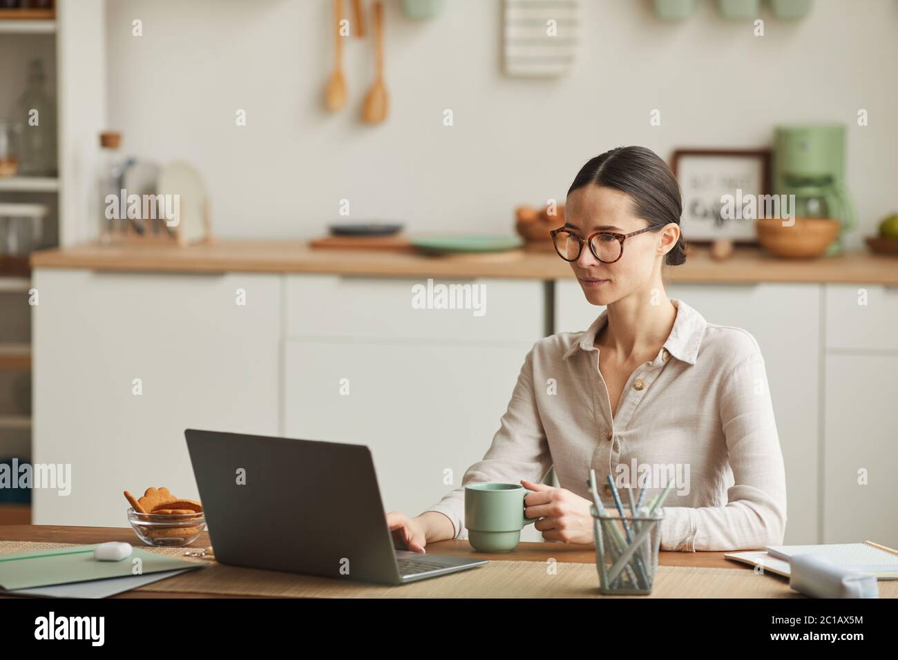 Portrait of elegant young woman drinking coffee while using laptop at cozy home office workplace, copy space Stock Photo
