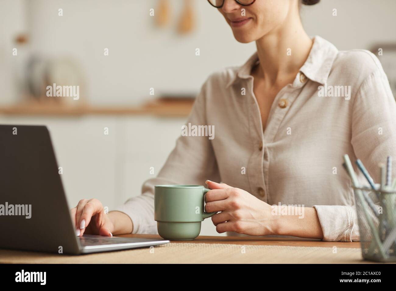 Cropped portrait of smiling young woman drinking coffee while using laptop at cozy home office workplace, copy space Stock Photo