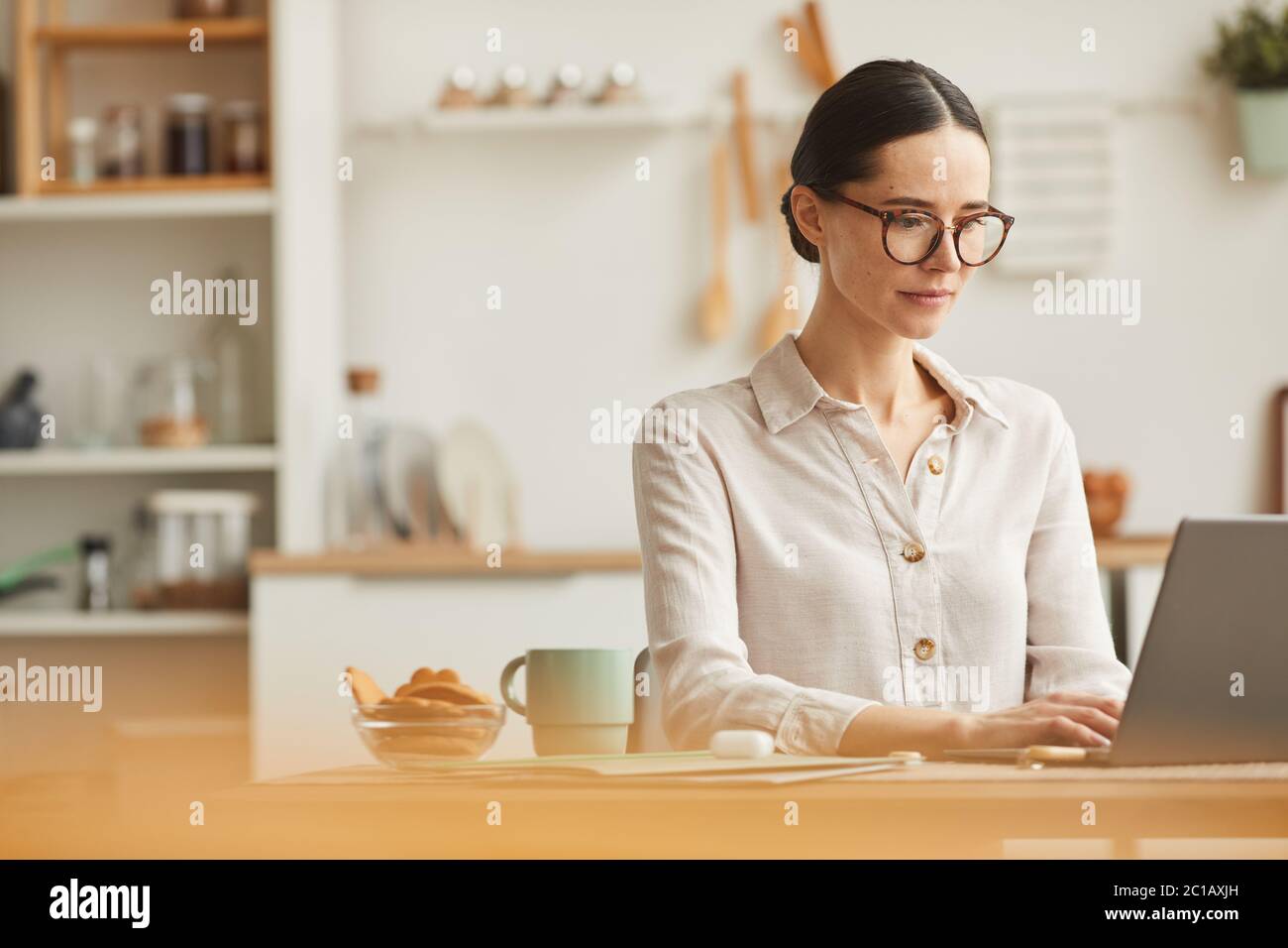 Warm-toned portrait of elegant young woman wearing glasses while using laptop at cozy home office workplace, copy space Stock Photo