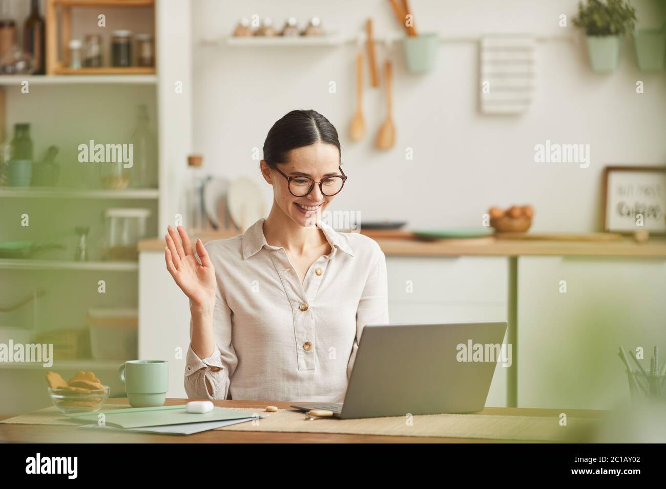 Warm-toned portrait of smiling young woman waving at camera during online meeting while working from home, copy space Stock Photo