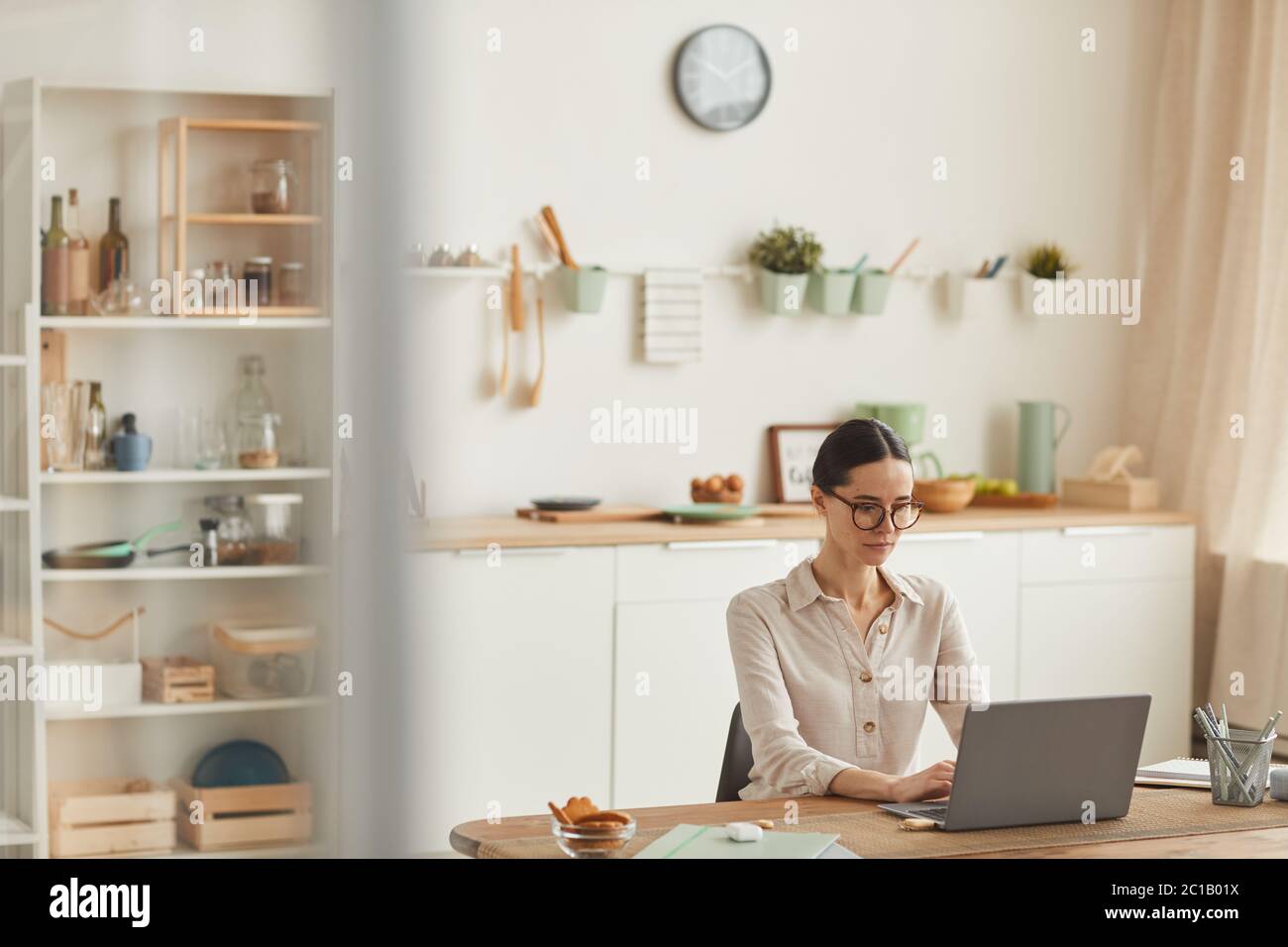 Warm-toned wide angle portrait of elegant young woman wearing glasses while using laptop at cozy home office workplace, copy space Stock Photo