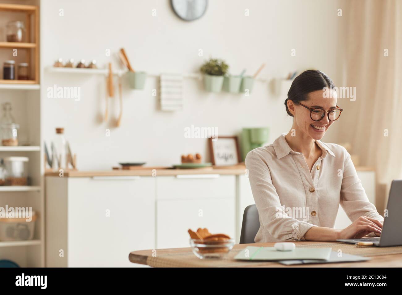 Warm-toned portrait of smiling young woman using laptop while sitting at cozy home office workplace, copy space Stock Photo