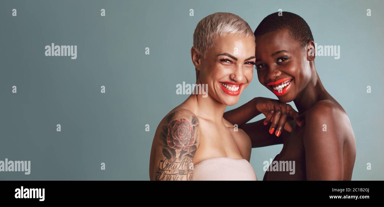 Two beautiful women standing together against gray background. Female models of different ethnicities with colorful makeup looking at camera and smili Stock Photo