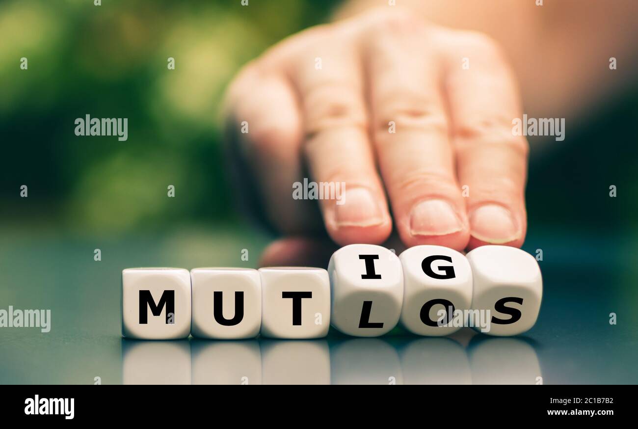 Hand turns dice and changes the German word 'mutlos' ('discouraged') to 'mutig' ('courage'). Stock Photo