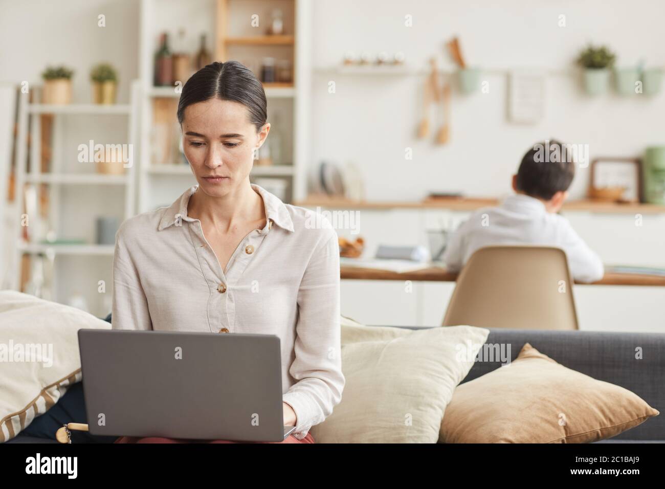 Warm-toned portrait of elegant young woman using laptop at home with her son studying in background, copy space Stock Photo