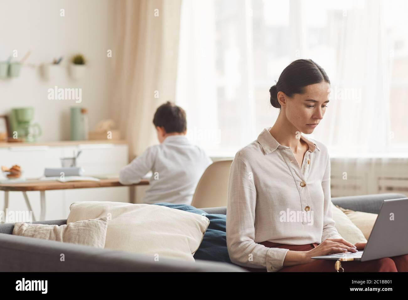 Warm-toned portrait of elegant young woman using laptop while working at home with her son studying in background, copy space Stock Photo