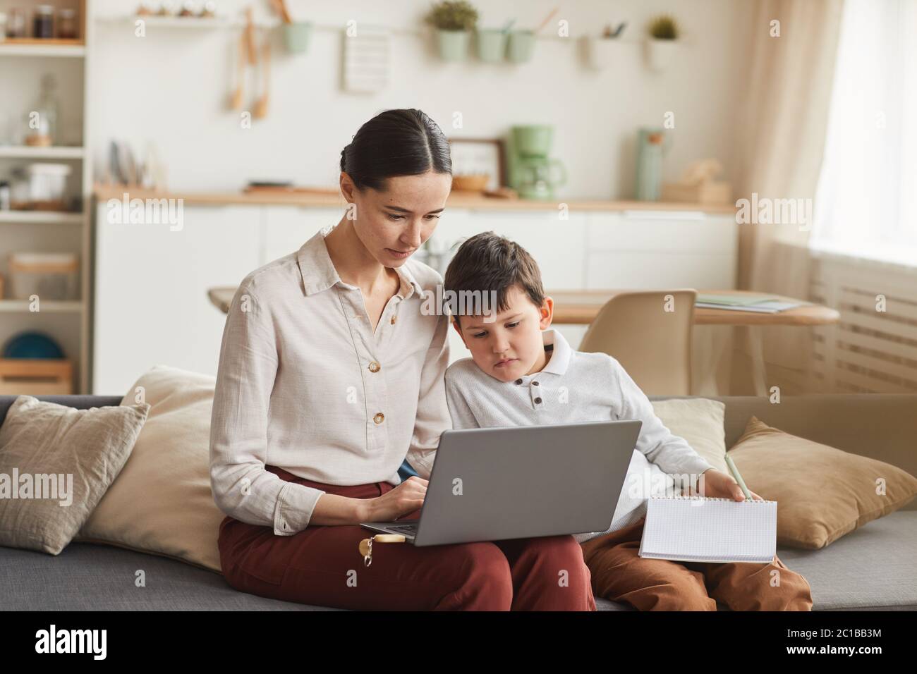 Warm-toned portrait of elegant young mother and son using laptop while studying at home in cozy interior, copy space Stock Photo