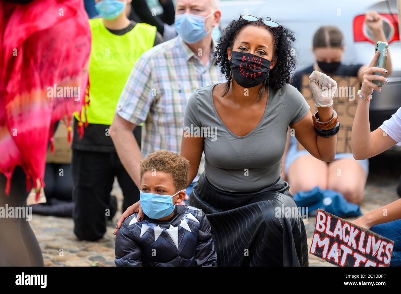 Richmond, North Yorkshire, UK - June 14, 2020: A woman wears a Black Lives Matter PPE Face Mask and salutes while holding her son at a BLM protest in Stock Photo
