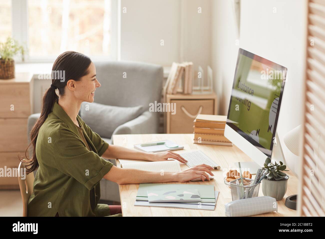 Warm-toned side view portrait of smiling young woman using computer while studying online at home, copy space Stock Photo