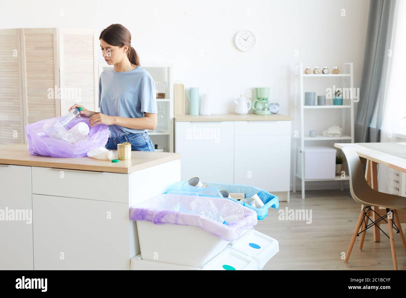 Young woman standing near the kitchen table and putting the garbage into the packet in the kitchen at home Stock Photo