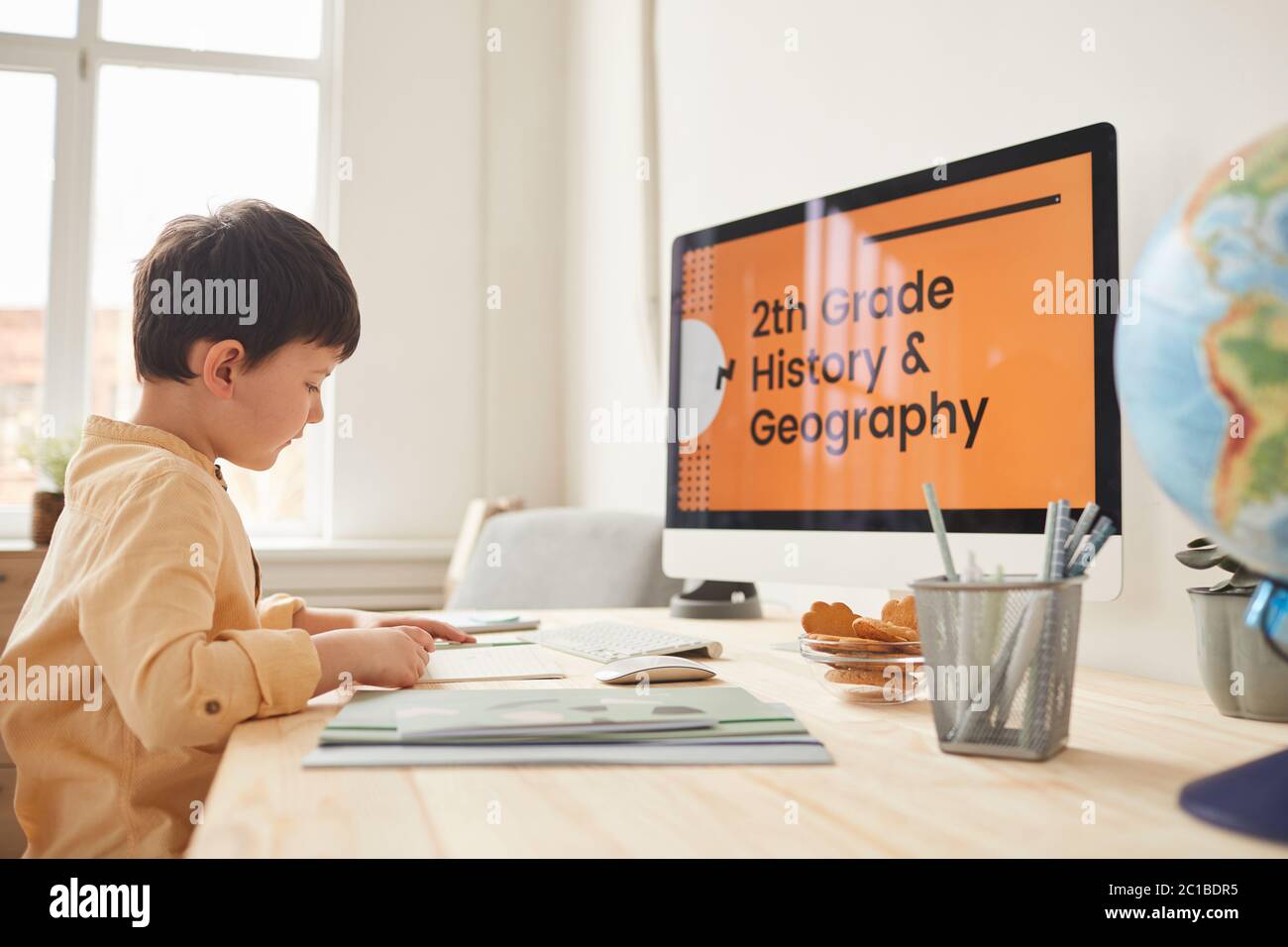 Warm-toned side view portrait of cute little boy doing homework while sitting at desk by computer with online school website, copy space Stock Photo
