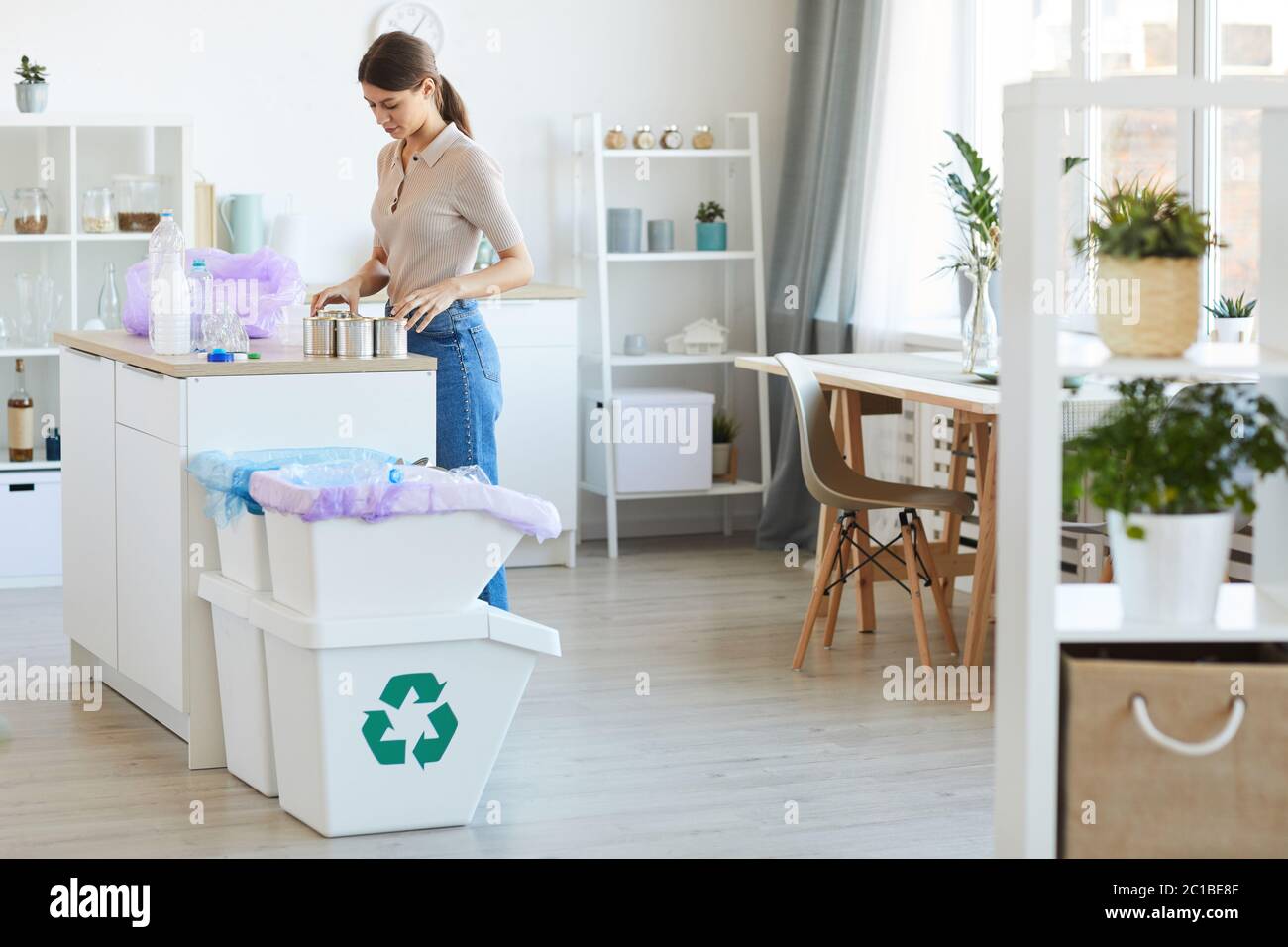 Young woman standing at the kitchen table and recycling plastic bottles and can Stock Photo