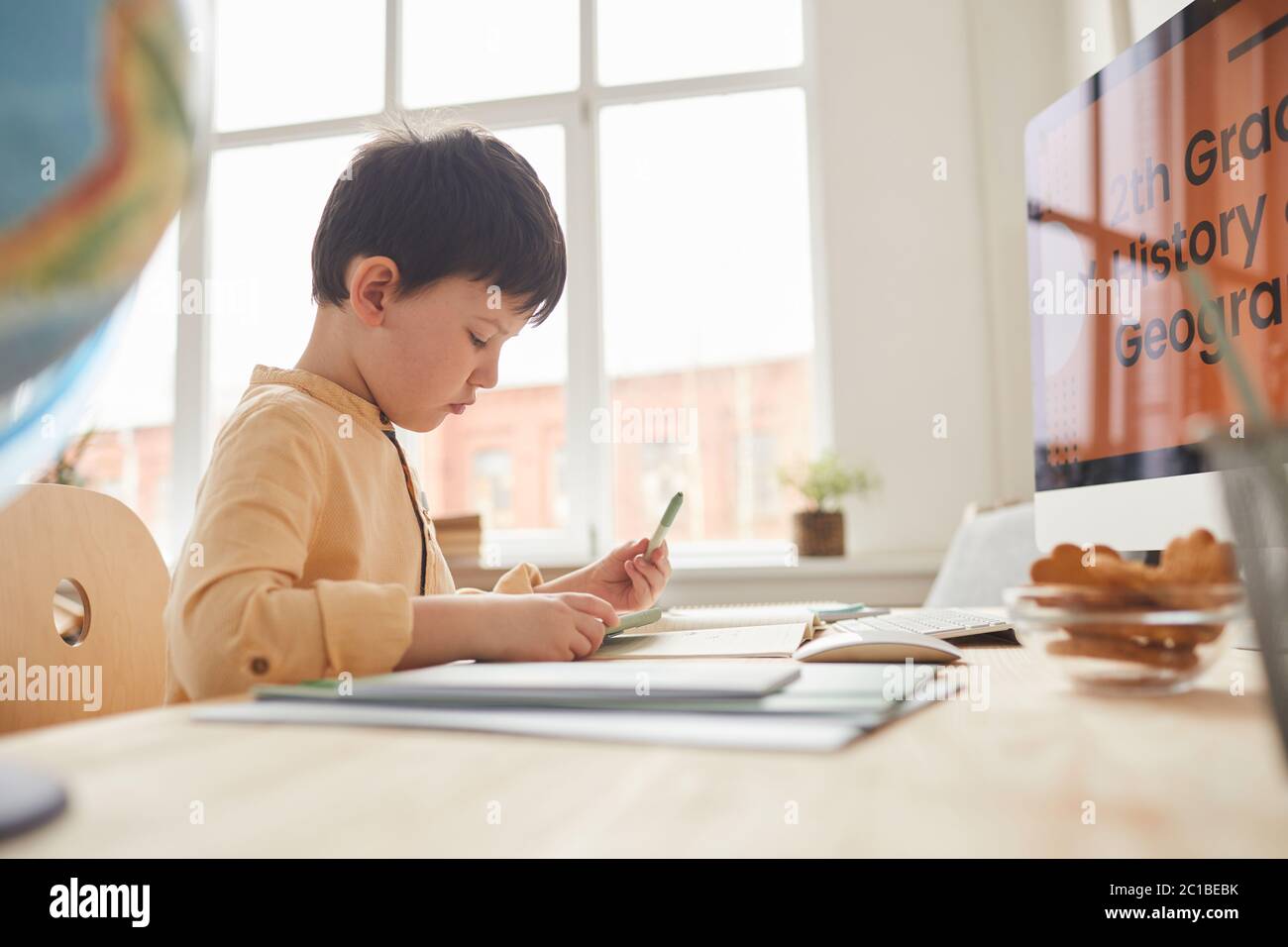 Sde view portrait of cute little boy doing homework while sitting at desk by computer with online school website, copy space Stock Photo