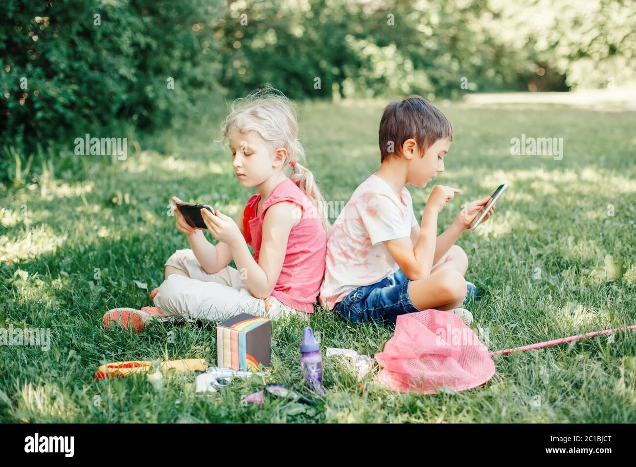 Boy and girl play games on smartphones outdoor. Kids digital gadget screen addiction. Children friends playing online sitting on grass in park. Stock Photo