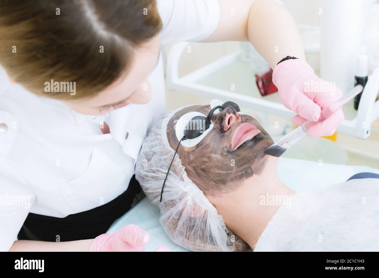 The cosmetologist in pink gloves with a brush applies a carbon mask for peeling on the face of a young girl in a cosmetology roo Stock Photo