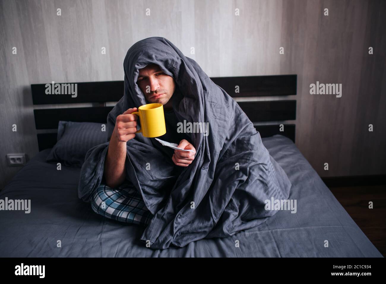 The man got sick. Lies in bed and suffers. He has fever, flu, colds and snot. He covered himself with a blanket and holds a napkin and hot tea Stock Photo