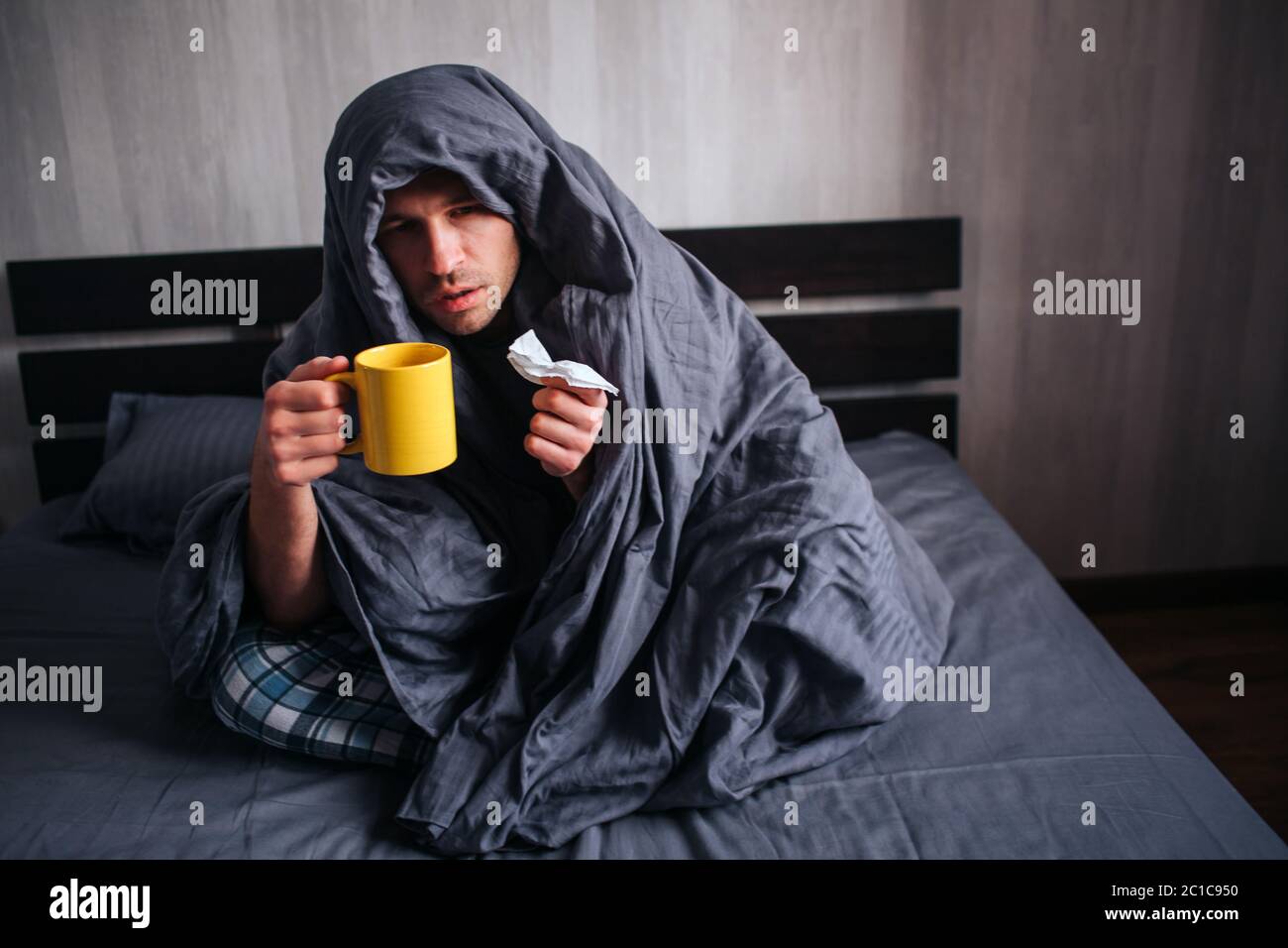 He covered himself with a blanket and holds a napkin and hot tea. The man got sick. Lies in bed and suffers. He has fever, flu, colds and snot Stock Photo
