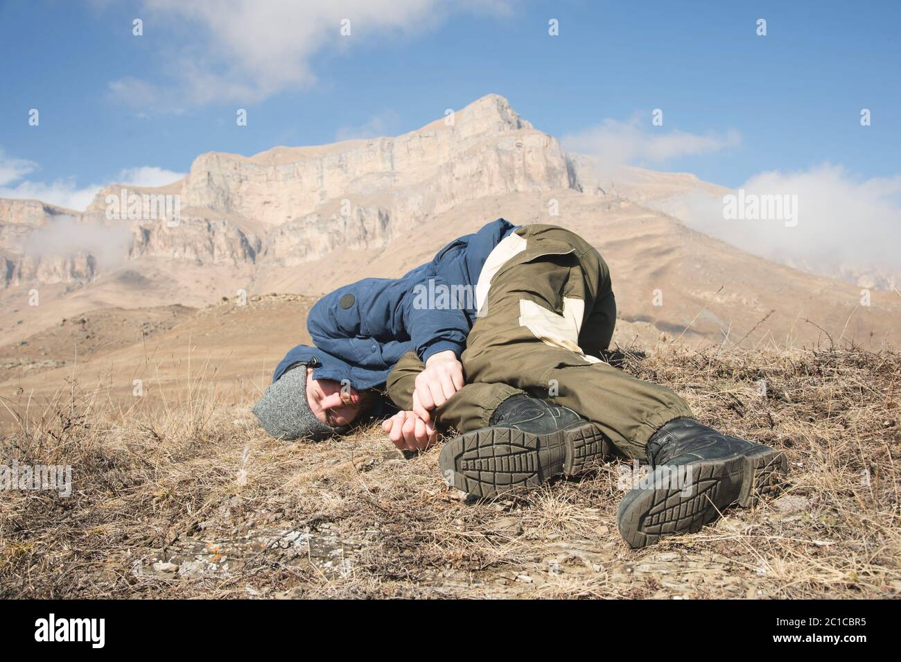 A tourist with a hat and a beard lies on the ground and feels bad in the mountains. A person who has lost consciousness. Danger Stock Photo