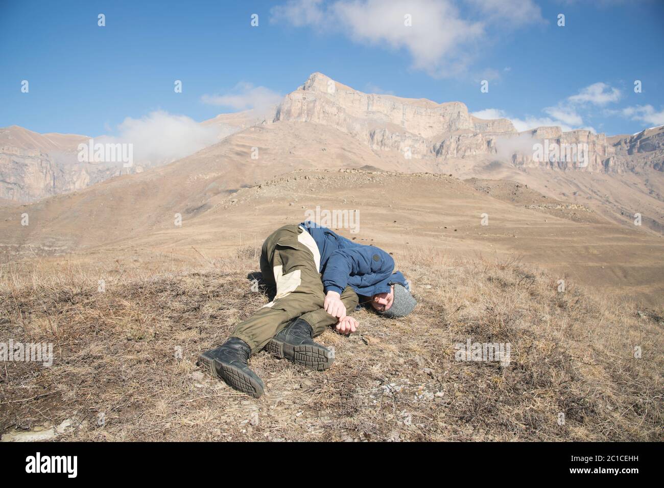 A tourist with a hat and a beard lies on the ground and feels bad in the mountains. A person who has lost consciousness. Danger - Stock Image