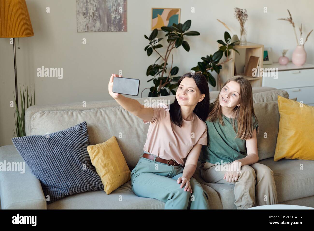 Smiling mother sitting on sofa in living room and taking selfie with daughter on smartphone Stock Photo