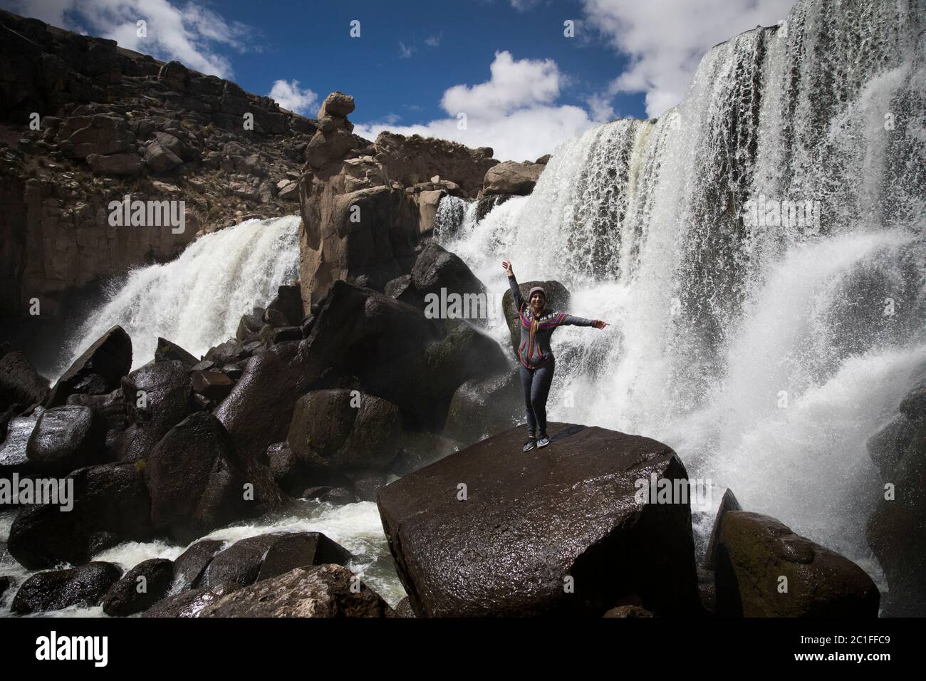 young woman standing with her open arms on a rock near a big waterfall, clouds and blue sky Stock Photo