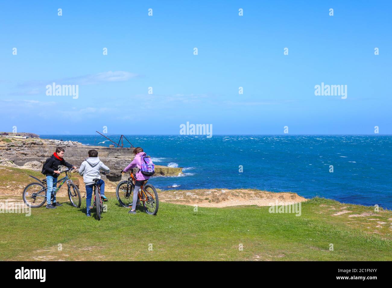 Isle of Portland, Dorset / UK - May 23, 2020: Three teenager with mountain bike on the cliff edge with blue sky and horizon over the water. Stock Photo