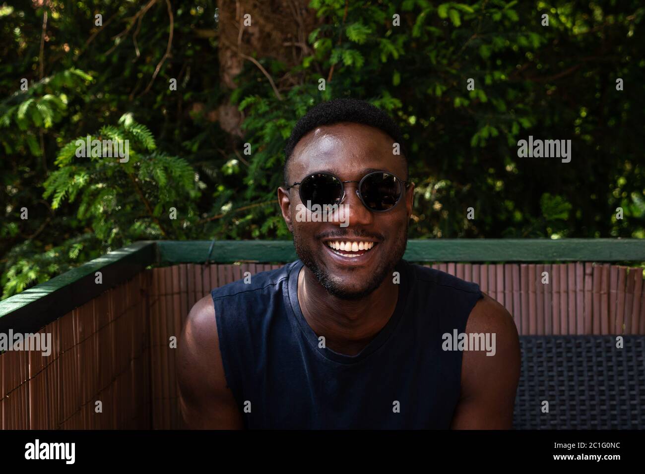 Young black man in sunglasses seated looking at camera and laughing. Medium shot. Stock Photo