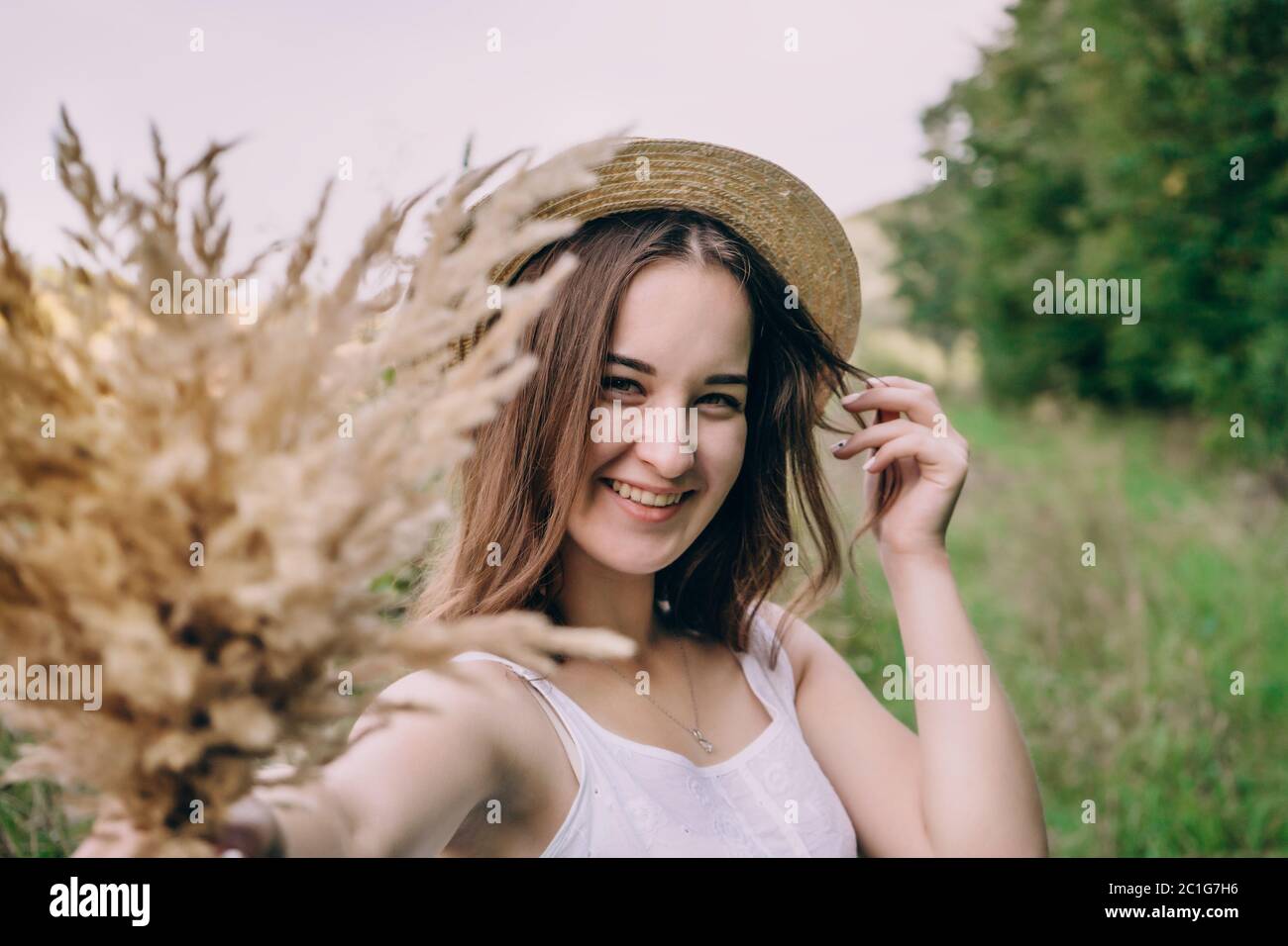 Happy young woman in a white dress in a straw hat. Smiling girl with a bouquet of dried flowers. Girl walks in a green park on a spring day Stock Photo